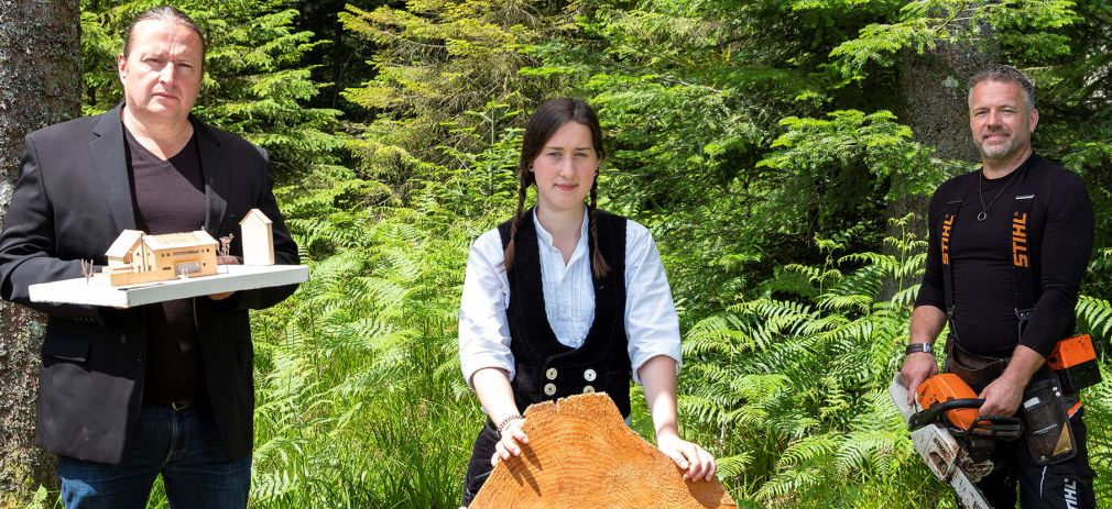 Flyerbild Faszination Weißtanne: Vom Baum zum Haus Drei Personen im Wald mit Modellentwurf, Holzscheibe und Motorsäge